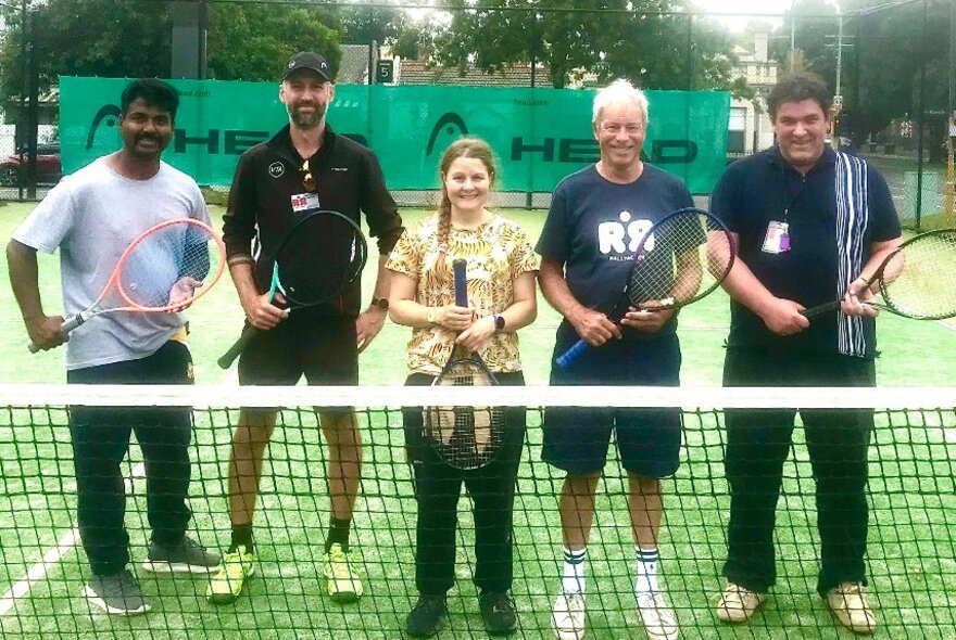 Five people standing at a net on a tennis court, racquets on hand.
