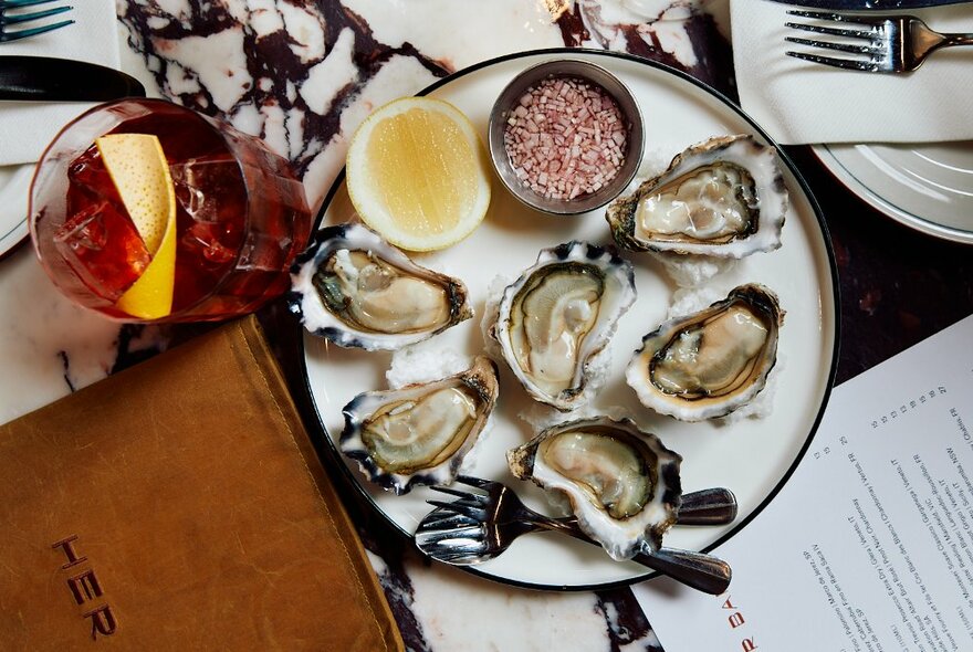 Overhead view of a plate of freshly shucked oysters served with half a lemon, a drink, a menu and cutlery nearby.
