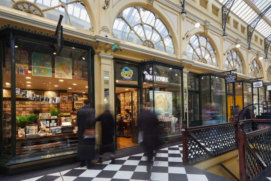 External view of shopfront in Royal Arcade with arched windows above doorway, black and white tiled arcade floor and shoppers strolling past.