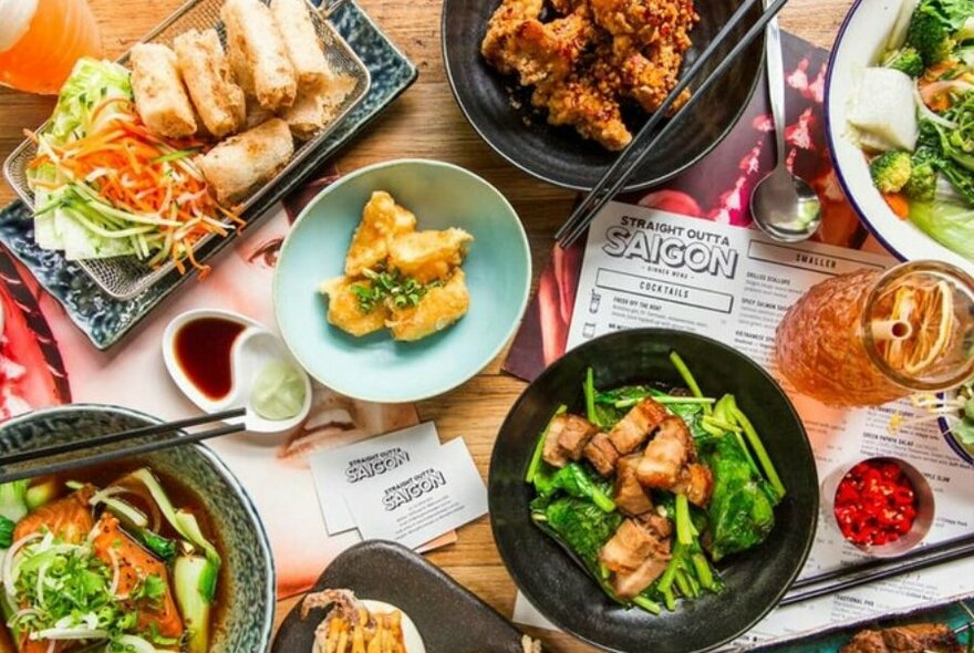 A table with a plate of spring rolls, a plate of fried chicken, a plate of dumplings, a plate of green vegetables and pork belly, a noodle soup and a cocktail. 