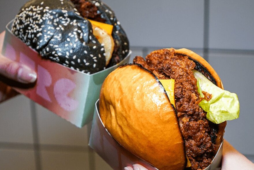 Two burgers held side-by-side. One is filled with fried chicken, the other with beef and a charcoal bun. 