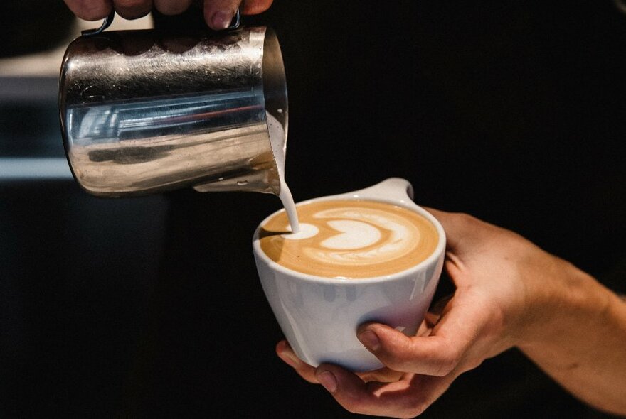 Milk being poured from a silver jug to create latte art on a coffee in a white cup. 