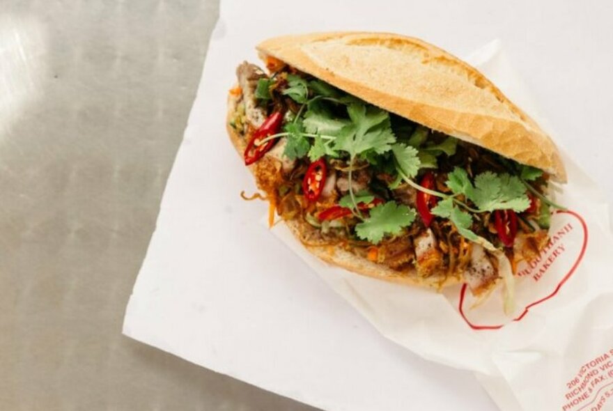 A banh mi filled with coriander, pork and chilli on a white paper bag.