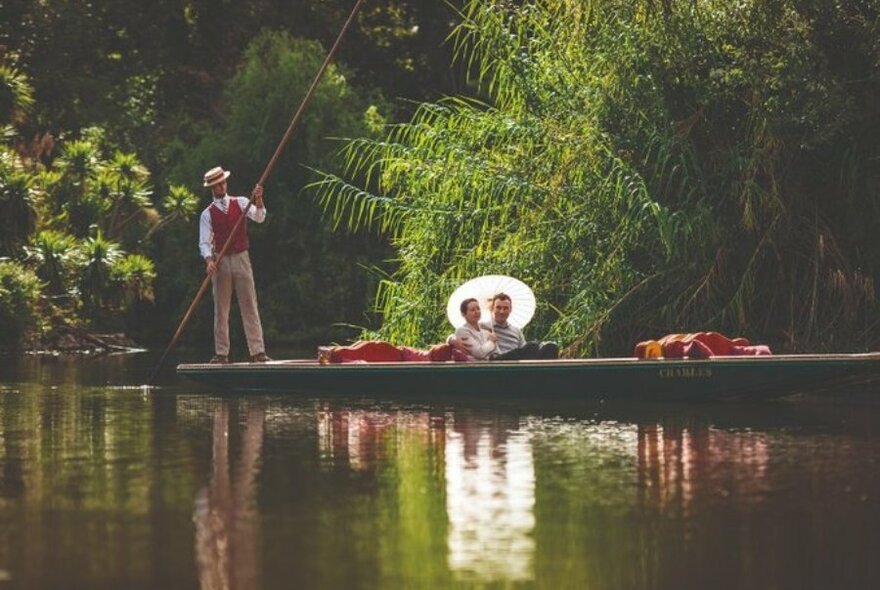 A couple on a boat surrounded by trees as a man paddles standing up.