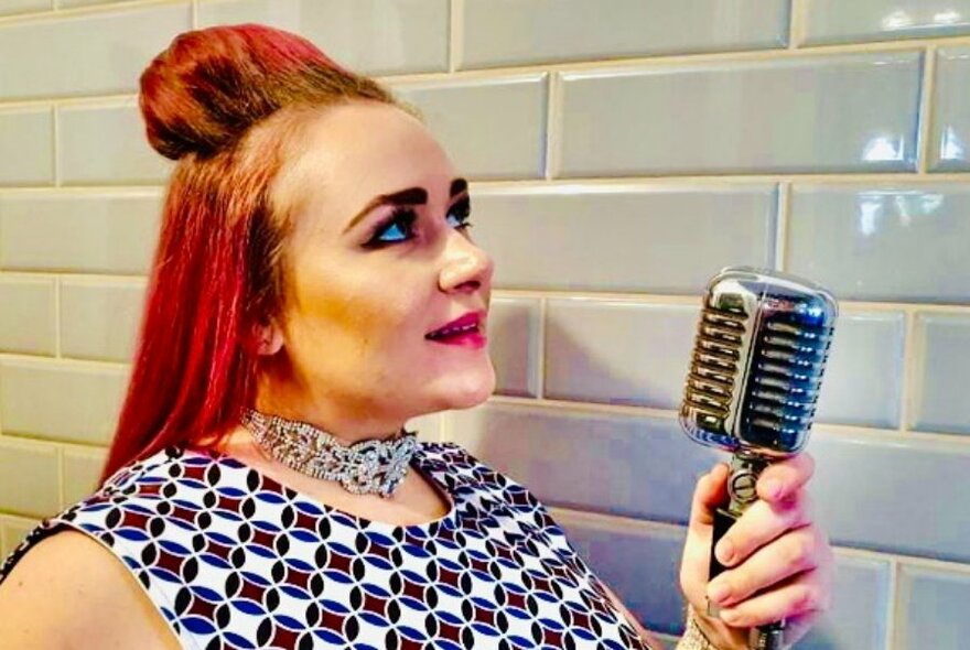 A woman with bright red hair and a large silver choker holds a retro microphone in front of a tiled background. 