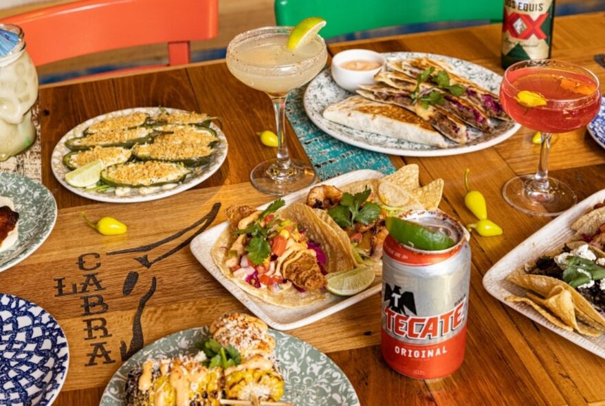 Selection of Mexican food on individual plates on a wooden tabletop in a restaurant, with two cocktails and two cans of beer also on the table.