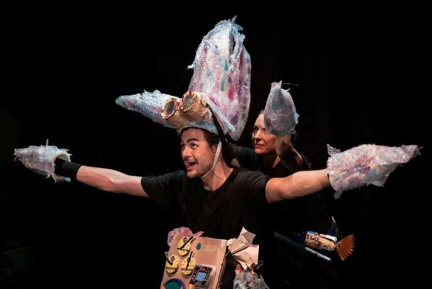 Two performers wearing garments made from recycled trash, posed with outstretched arms against a black background.
