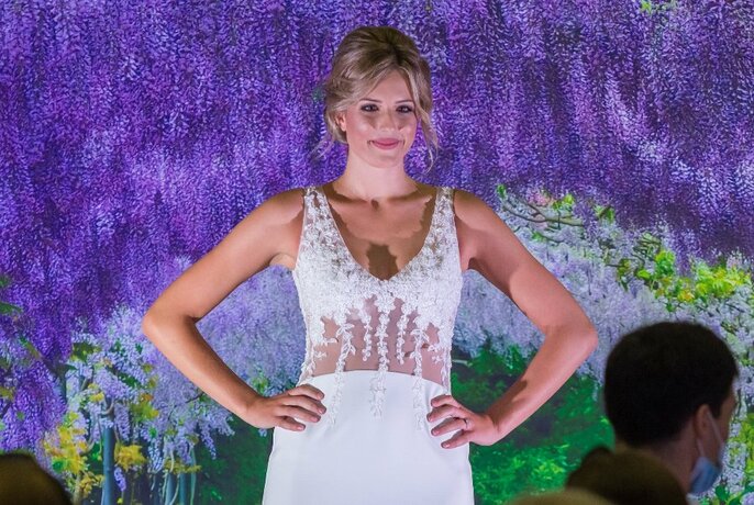 Model smiling with hands on hips wearing sleeveless white dress in front of floral background.