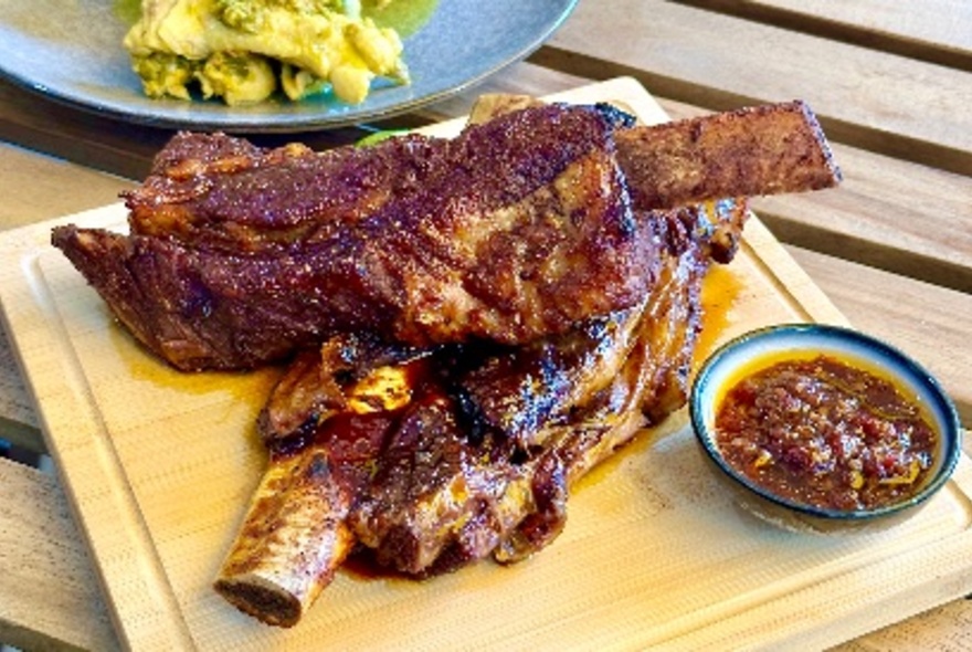 Two large beef ribs served on a a wooden tray with a dipping sauce.