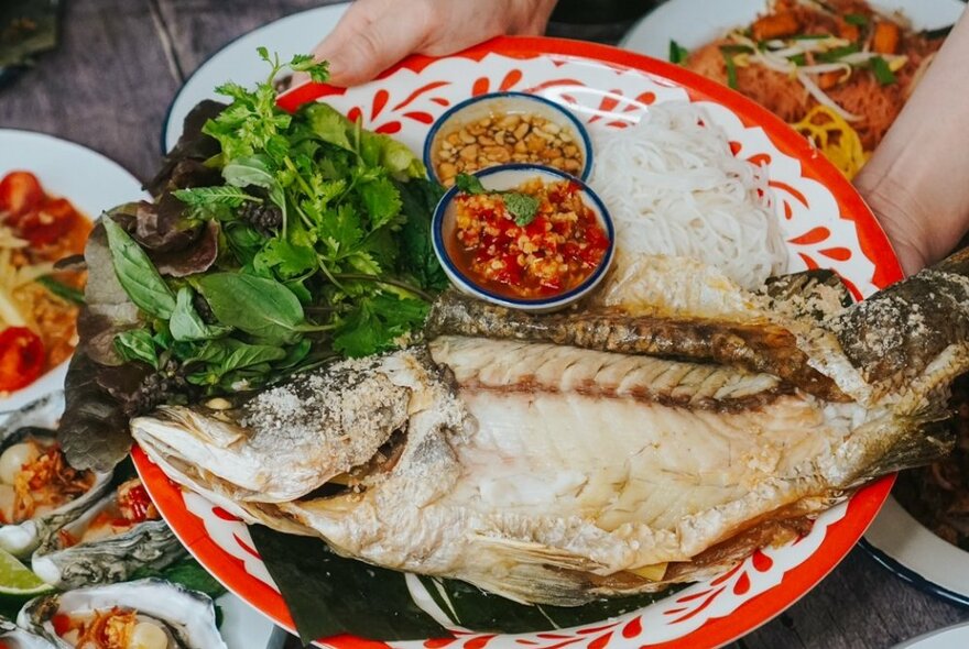 A whole baked fish on a plate with condiments and greens. 