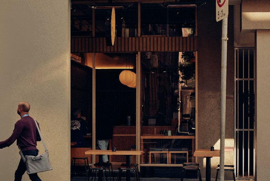 A man with a satchel walking past a restaurant with Japanese-style architecture and interior design.