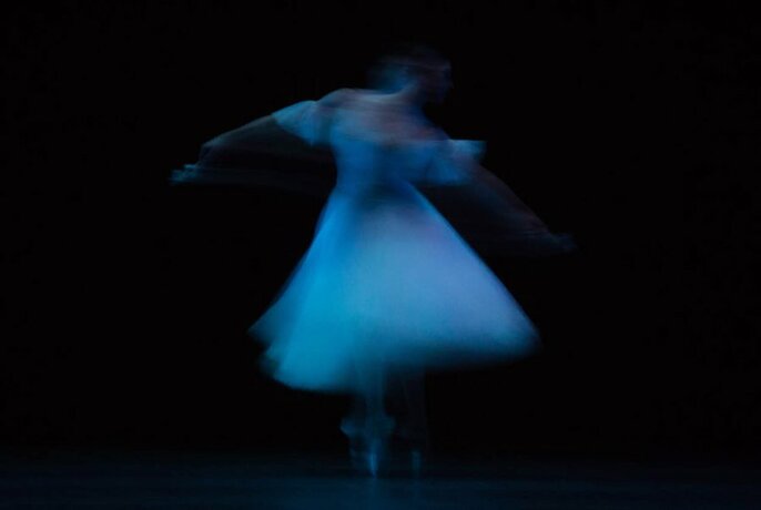 A ballet dancer wearing a dress, twirling on a stage on pointe shoes, blurred in motion against a black background.