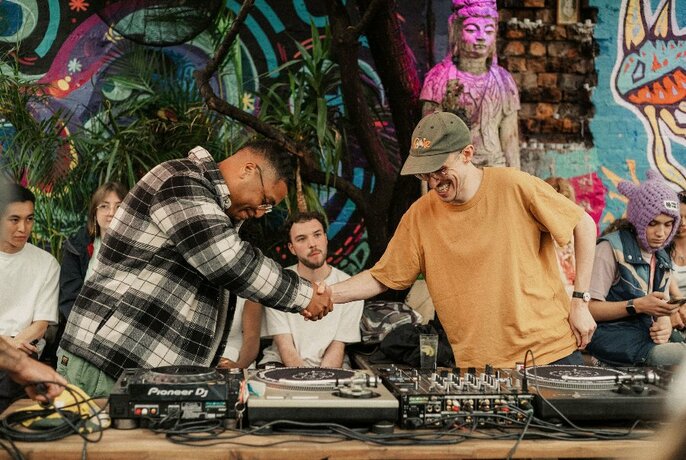 Two people shaking hands in a friendly fashion over a DJ mixing desk of turntables.