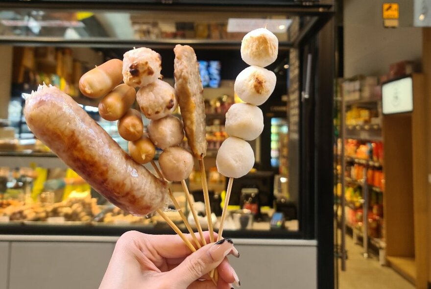 A hand holding up skewers of cooked hot food outside the front of a mini supermarket