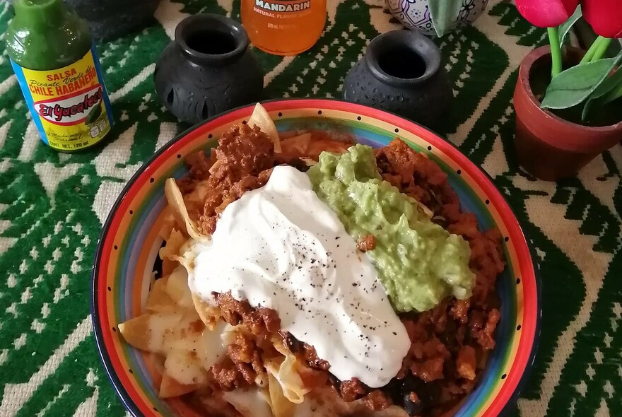 A plate of nachos topped with sour cream and guacamole and drinks in the background all on a green tablecloth.
