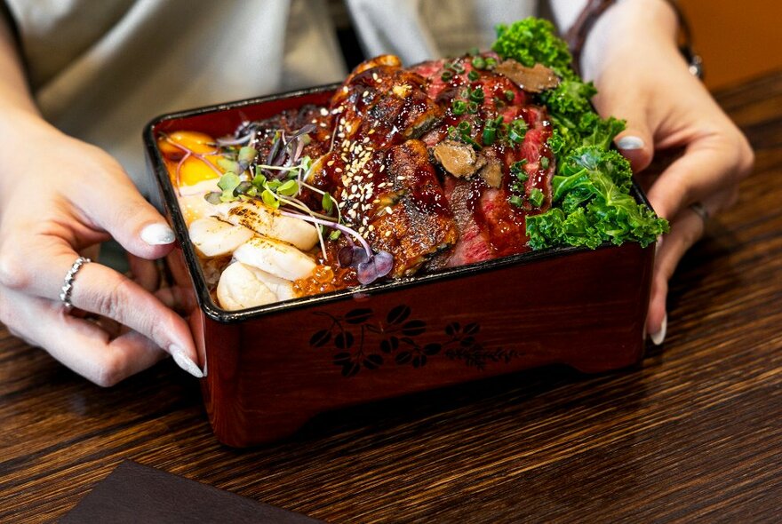 Hands holding a Japanese meal in a box featuring wagyu beef. 