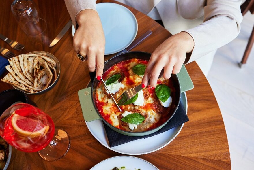 Cutting into a large duck lasagne served in a bowl, there is also bread and a cocktail on the table 