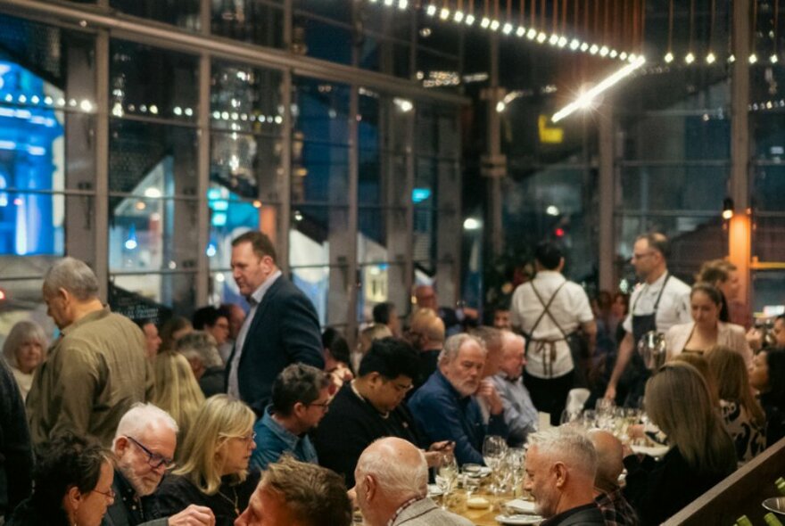 Dining patrons eating and drinking, seated at long tables inside a restaurant, with large windows surrounding them.