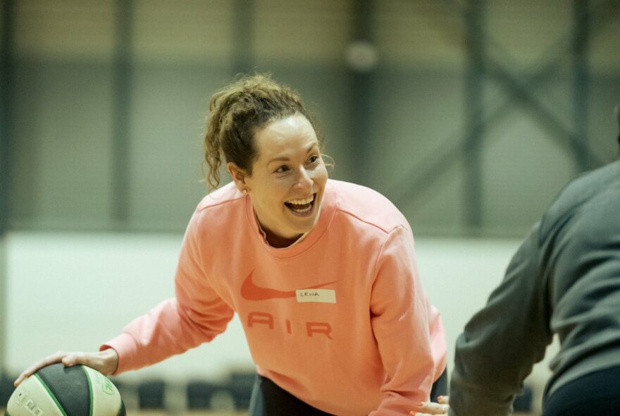 A smiling woman in a peach jumper, bouncing a basketball inside a basketball stadium.