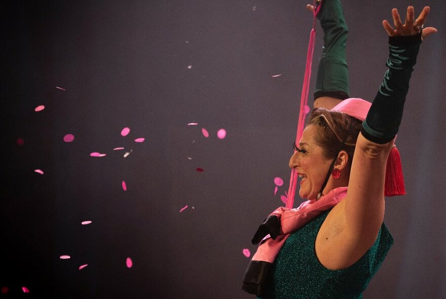 A side view of a woman performing with her hands in the air and pink confetti fluttering in front of her.