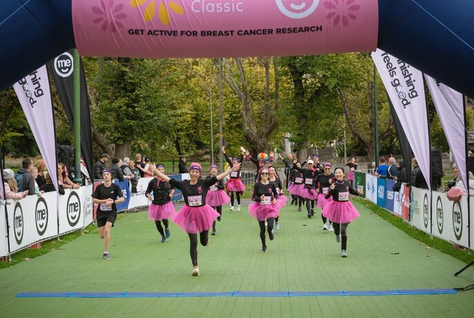 People in black, with pink tutus, running through large, blow-up banner.