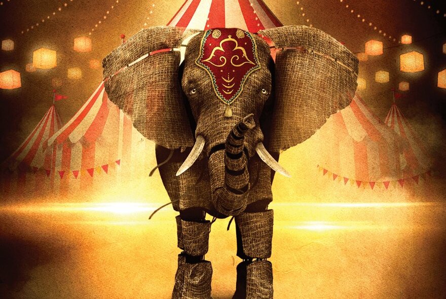 A large elephant puppet wearing a circus headdress with segmented legs, with a big top in the background and a spot light shining down.
