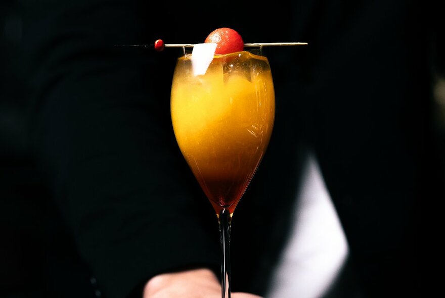 An orange coloured cocktail in a glass with a cherry garnish.