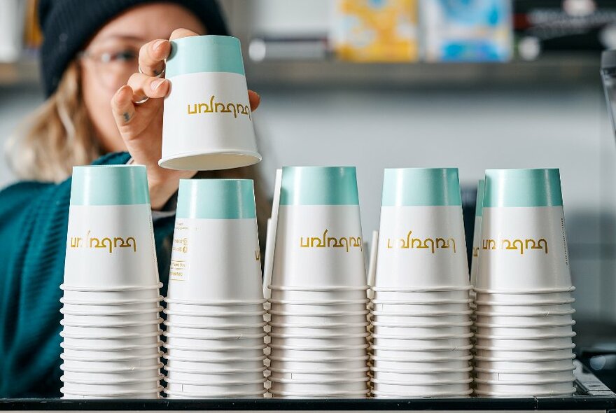 A barista takes a cup from a large stacked line-up of takeaway cups. 