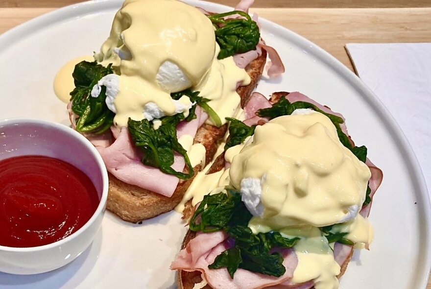 A breakfast plate of poached eggs, spinach, ham and hollandaise sauce on toast, with a side of tomato sauce.