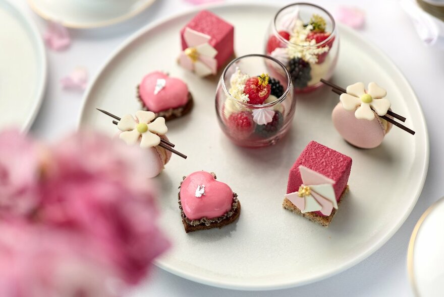A plate of dainty little petit fours, all in pink tones with a blurred pink flower in the foreground. 