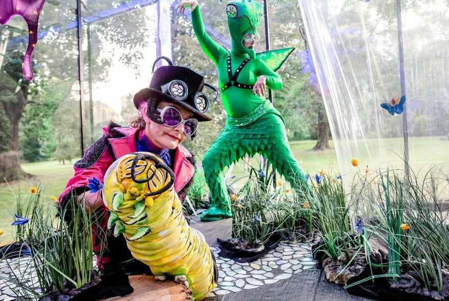 Performers dressed in costumes, one in a head to toe green insect costume, and another in a pink jacket and black top hat holding a yellow caterpillar puppet, in an outdoor setting with some plants in pots nearby. 