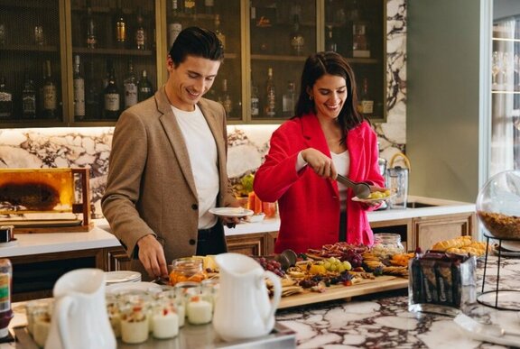 Two smiling people selecting food to put on their plates from an extensive breakfast buffet.