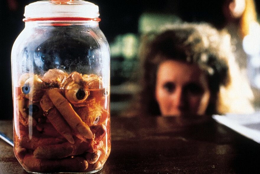 A clear glass jar of bloody severed fingers, and a woman's blurred and out of focus face in the background.