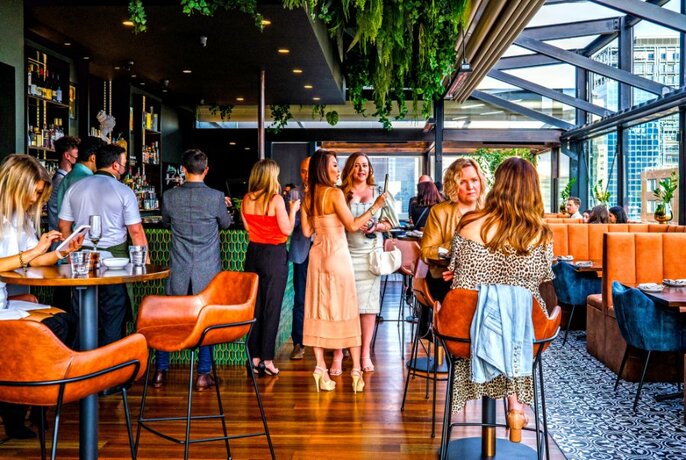 A busy indoor-outdoor rooftop bar with orange seating and greenery hanging from the roof.