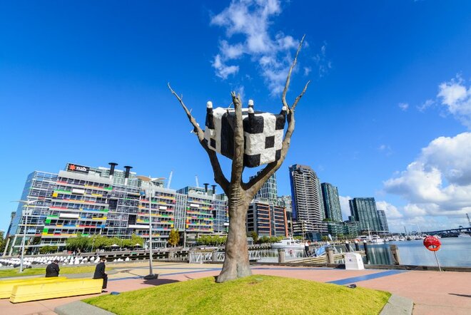 Large sculpture of an upside-down black and white cow in a barren tree, next to the waterfront and apartments.