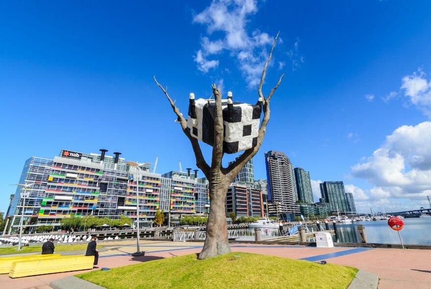 Large sculpture of an upside-down black and white cow in a barren tree, next to the waterfront and apartments.
