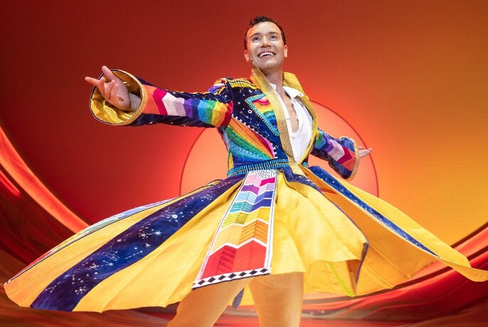 A performer twirling on a theatre stage, wearing a brightly coloured coat.