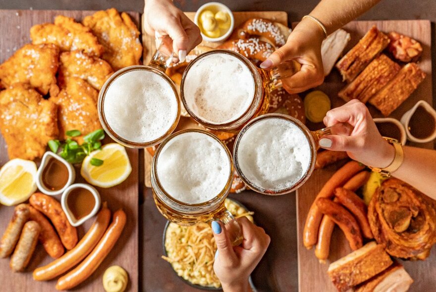 Plates of food including large pretzels, sausages, schnitzels, and chips on a table, with fours hands holding large steins of beer above the food on the table.
