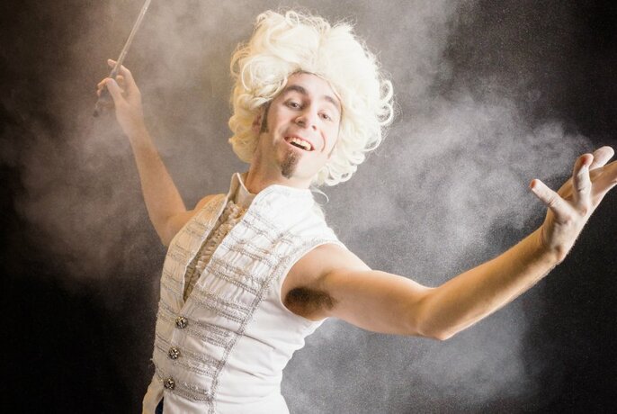 A person in a white curly wig and white vest holding a wand in a puff of smoke. 