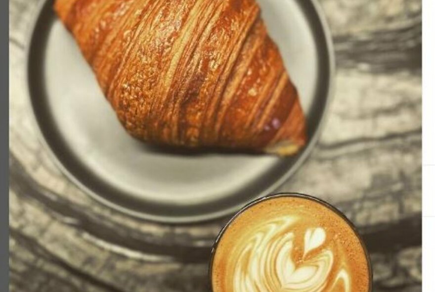A croissant on a grey plate and a coffee on a marble table seen from above.