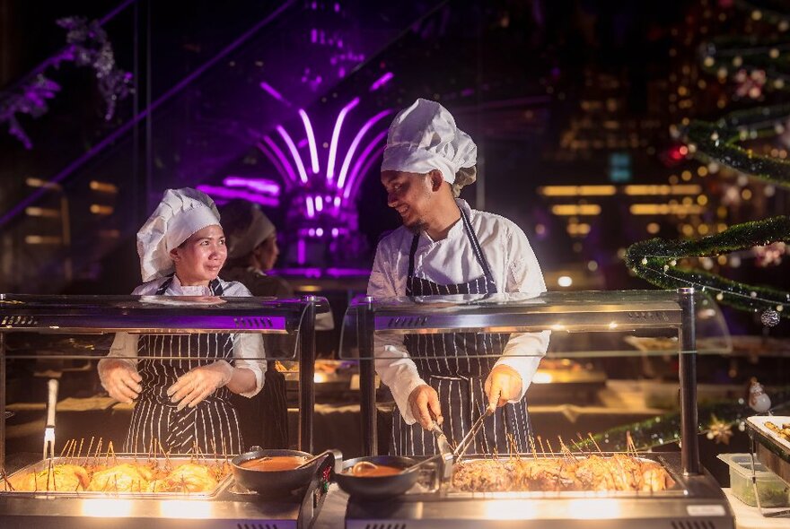 Two cooks in chef whites and hats and striped aprons standing behind an outdoor glassed food service area, with city buildings at night behind them.