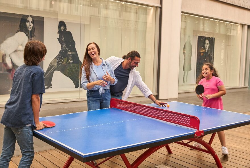 A family playing table tennis on a blue table, in a large open space within an indoor shopping precinct.