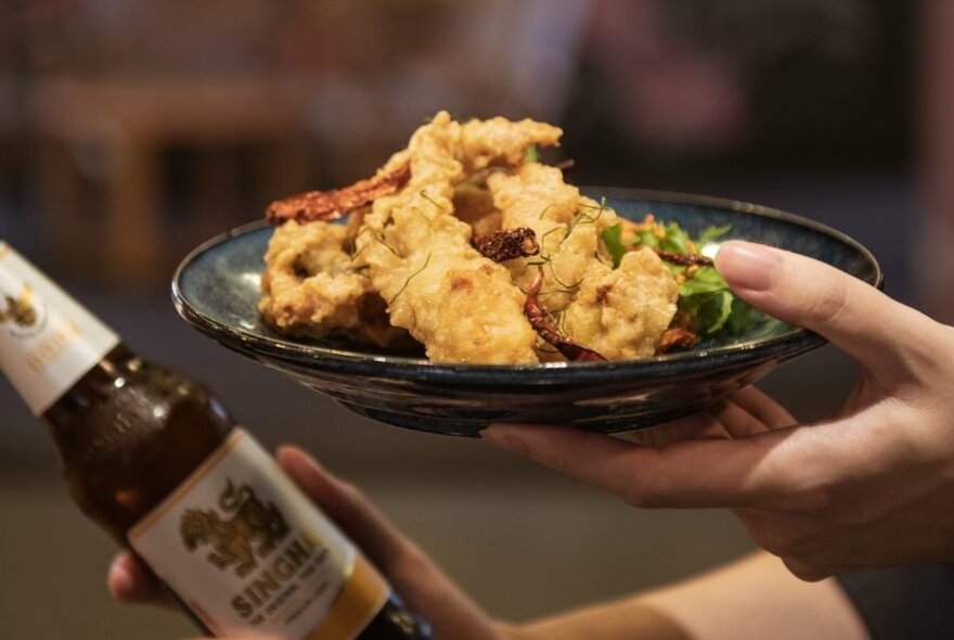 Hands holding a plate of fried Asian food and a bottle of beer.