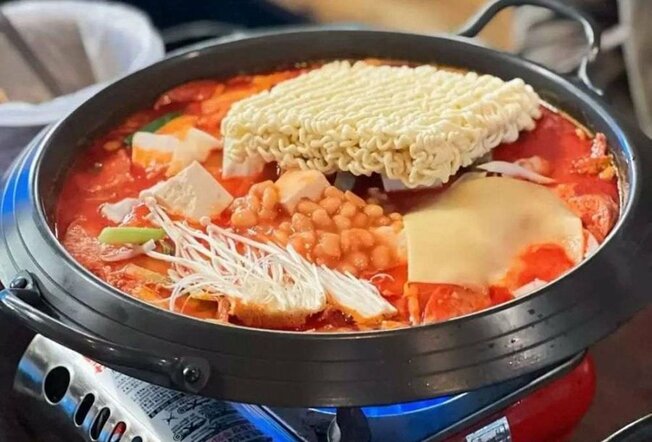 A black pot on a gas burner, filled with noodles, red broth, baked beans and vegetables