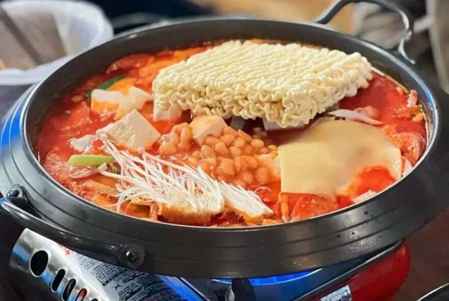 A black pot on a gas burner, filled with noodles, red broth, baked beans and vegetables