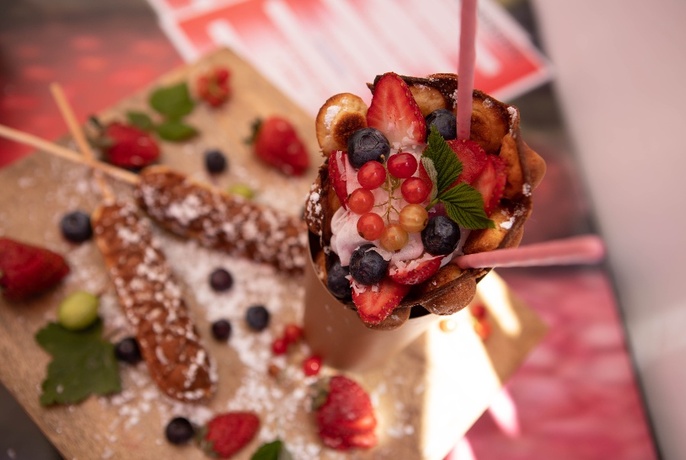 Close up of fruit-covered dessert, with fried goods on sticks, sprinkled with icing sugar on surface behind.
