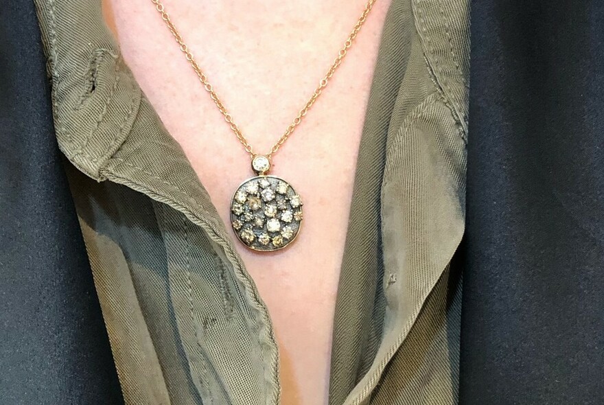 Circular, jewelled pendant hanging against pale skin, between olive green shirt and dark blazer.