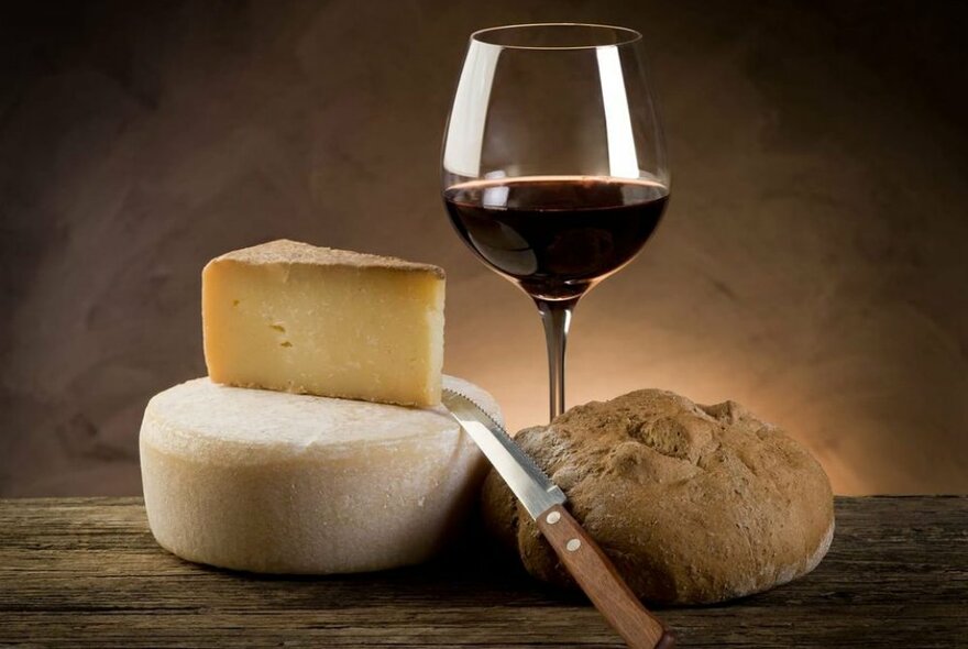 A glass of red wine next to a bread roll, knife and two cheeses.