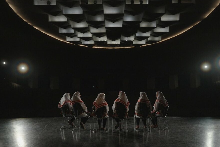Six people seated in a row of chairs, their backs to the viewer, in a large open dark space, like a theater stage.