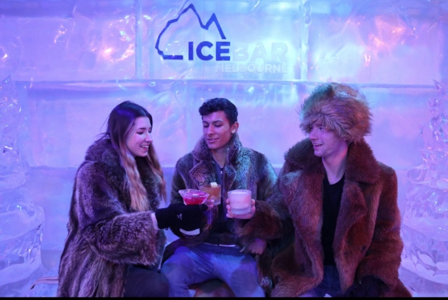 Three people wearing fur coats clinking glasses against an ice backdrop.